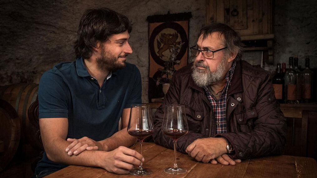Gian-Battista und Sohn Johann-Baptista sitzen an einem Holztisch im Keller und schauen sich bei einem Glas Rotwein in die Augen.