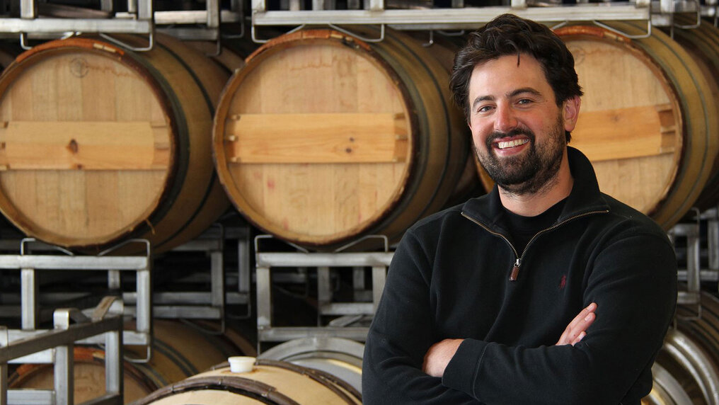 Matt Reynvaan lächelt in die Kamera. Im Hintergrund sind gestapelte Holzfässer zu sehen.