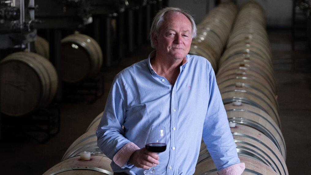 Peter Bright steht mit einem Glas Rotwein in der Hand vor Weinfässern im Keller