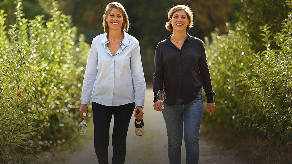 Adèle und Elsa Matrot schlendern mit Weingläser und einer Flasche Rotwein in den Händen durch einen Weinberg.