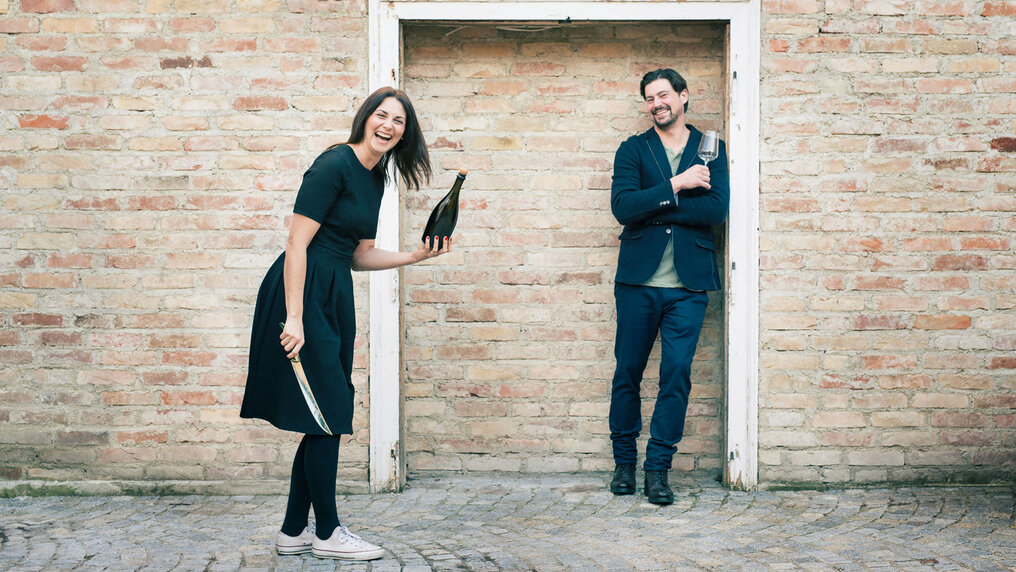 Marion is about to open a bottle of sparkling wine with a sabre, while Manfred leans against an old door frame set into the brick wall, a wine glass in his hand.