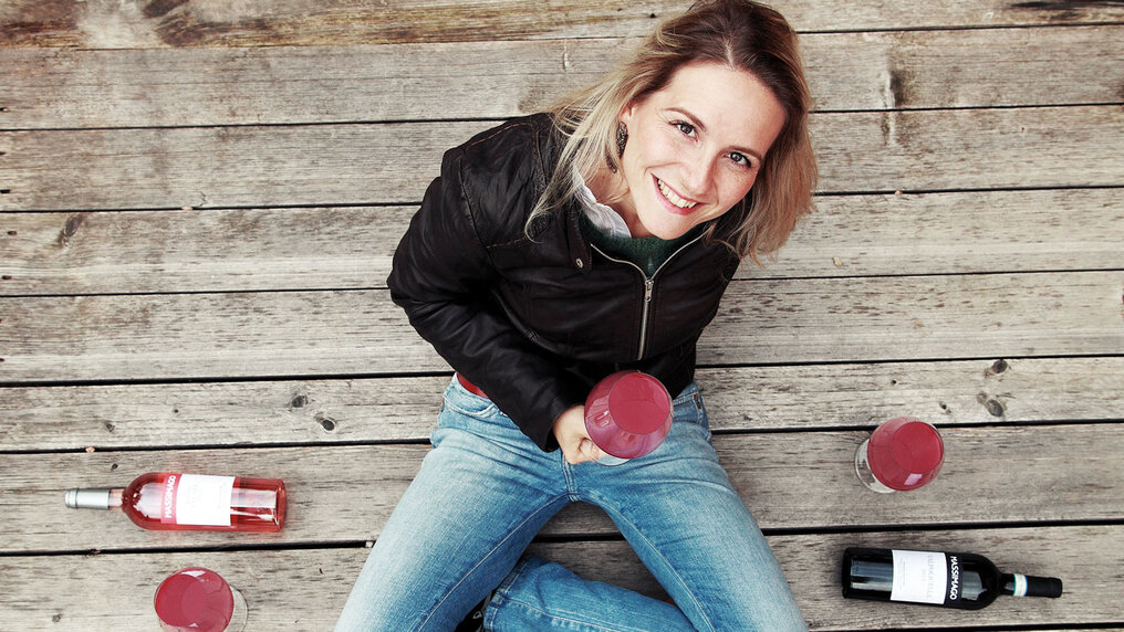 Camilla Rossi Chauvenet sits cross-legged on the wooden floor, a glass of red wine in her hand, looking up at the camera.