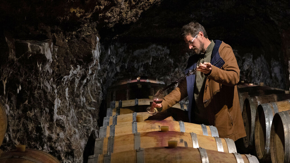 In his underground vaulted cellar made of tuff stone, Sylvain Dittière uses a pipette to take a sample of red wine from a wooden barrel and pours it into a glass.
